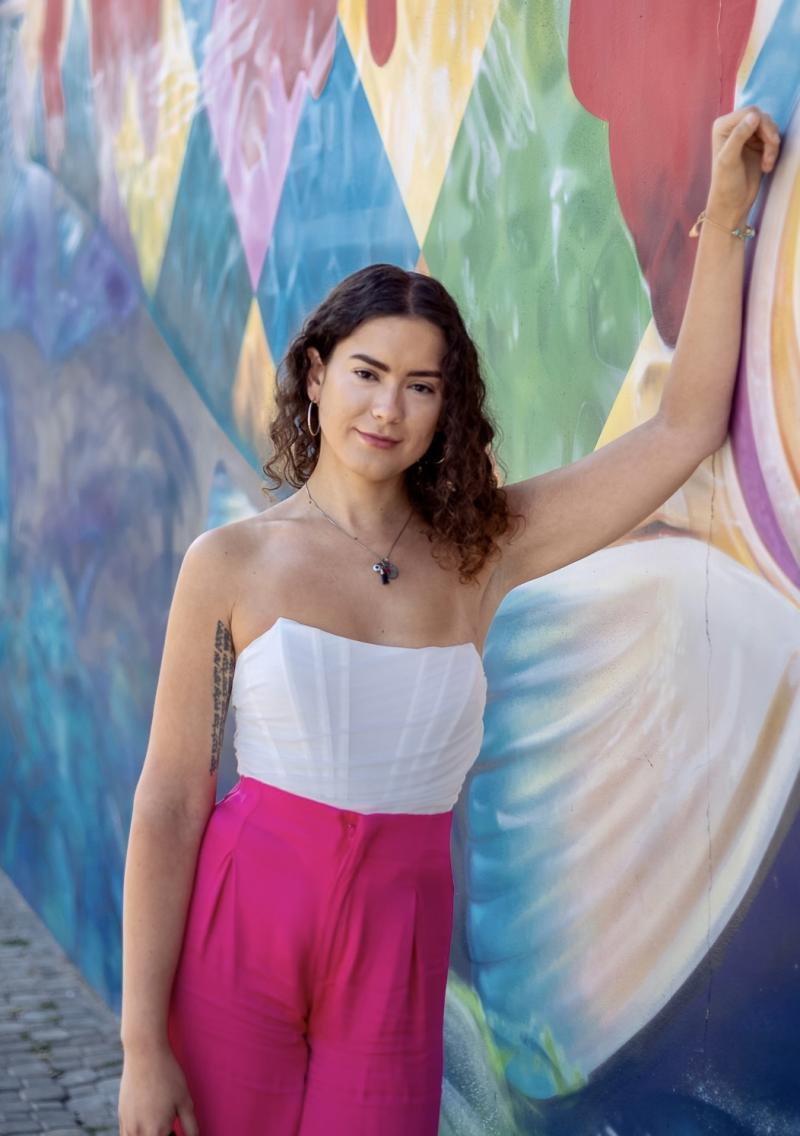 Headshot of LGBTQ CRC Directory, Vanessa Aviva, with a white top and pink pants next to a colored wall 