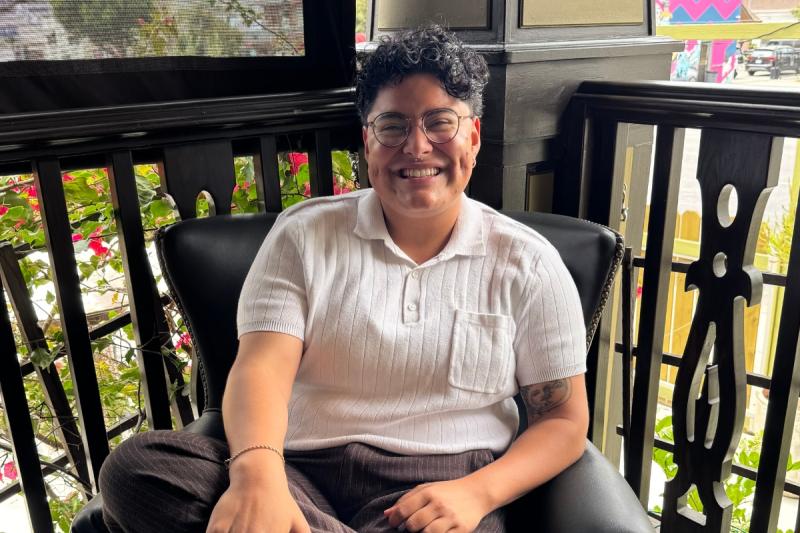 A photo of Gael Jerez-Urquia, LGBTQ CRC Program Coordinator. He is sitting on a chair in a corner in a white shirt. He has short black hair and glasses.