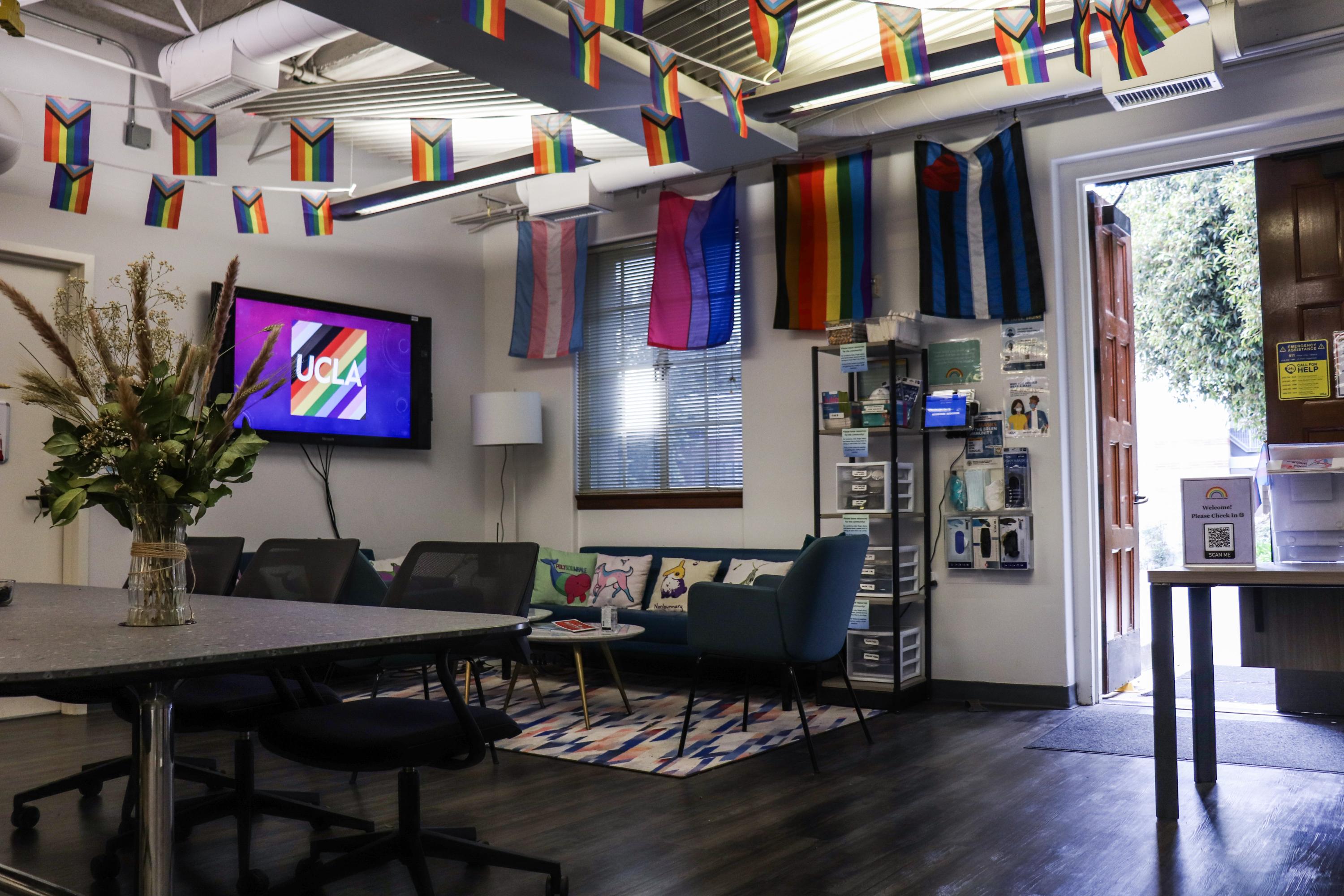 The inside lounge of the LGBTQ CRC with pride identity flags hanging from the walls a ceiling, a table, a couch, and the UCLA pride logo. 