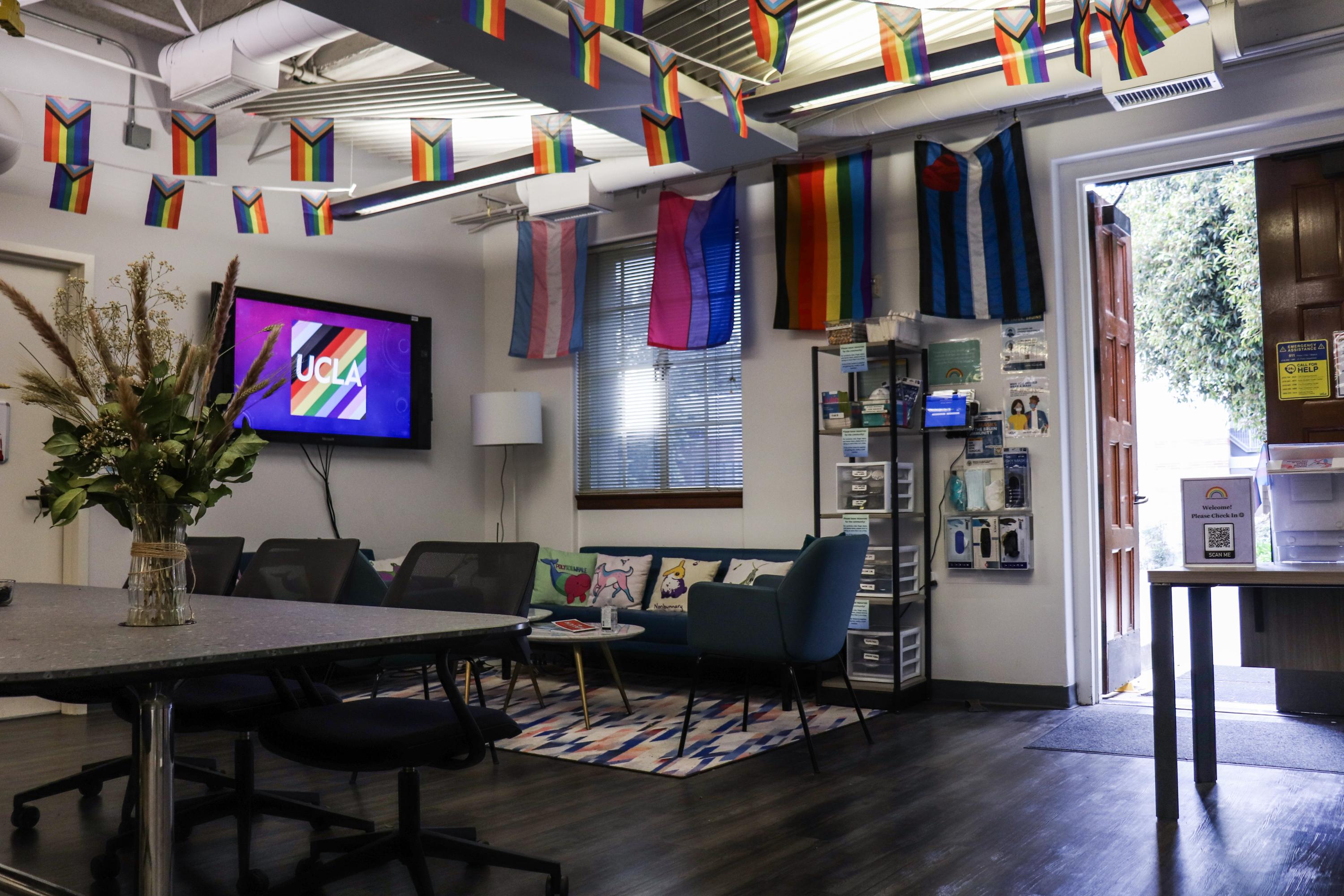 The inside lounge of the LGBTQ CRC with pride identity flags hanging from the walls a ceiling, a table, a couch, and the UCLA pride logo.