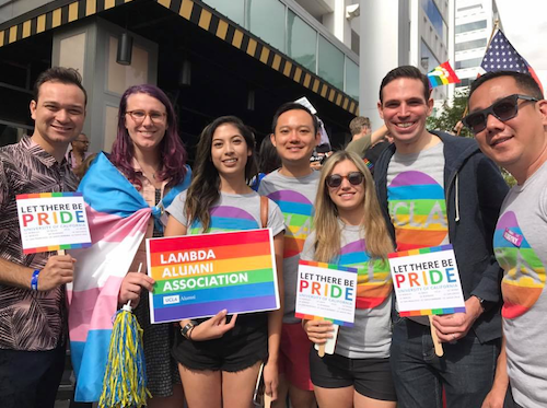 Members of Lambda Alumni stand together in t-shirts with rainbows on them, holding signs that say 'let there be Pride.' One alumn is wearing a transgender flag like a cape.