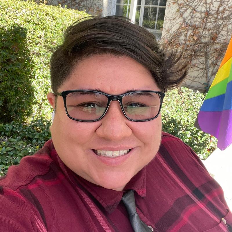 Minnie Esquivel Gopar is wearing a magenta plaid button up shirt with a black tie. They are smiling while wearing glasses and with short hair on the sides and a combed hair on top. They are in front of the green hedges and rainbow pride flag outside the LGBTQ CRC.