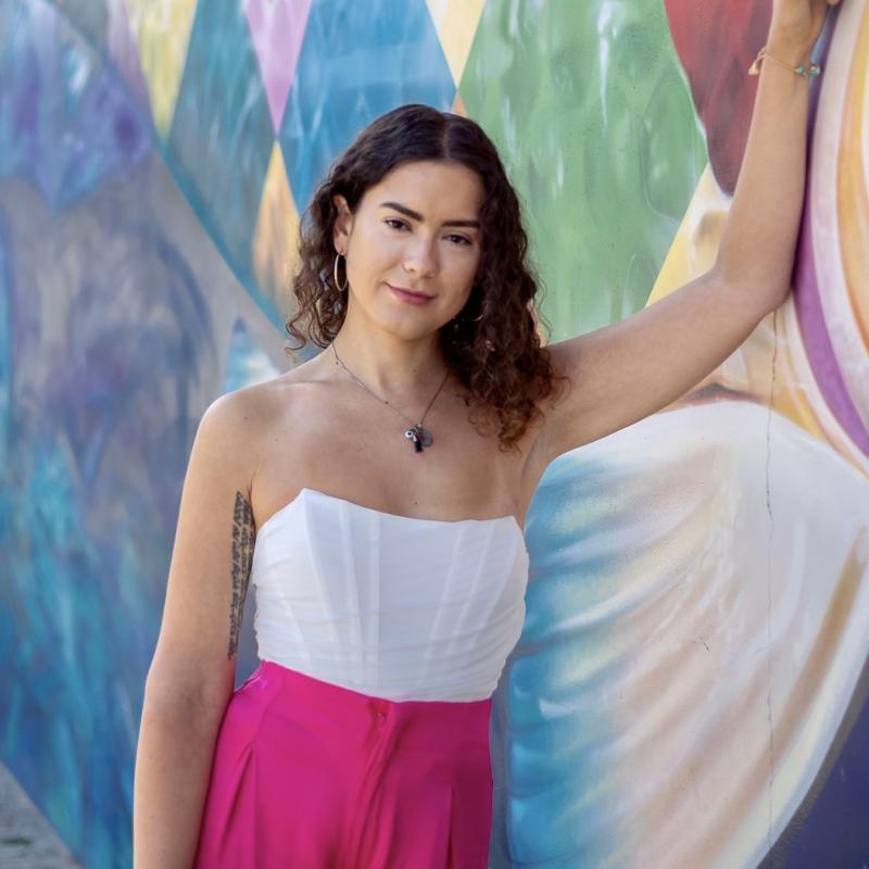 Headshot of LGBTQ CRC Directory, Vanessa Aviva, with a white top and pink pants next to a colored wall 