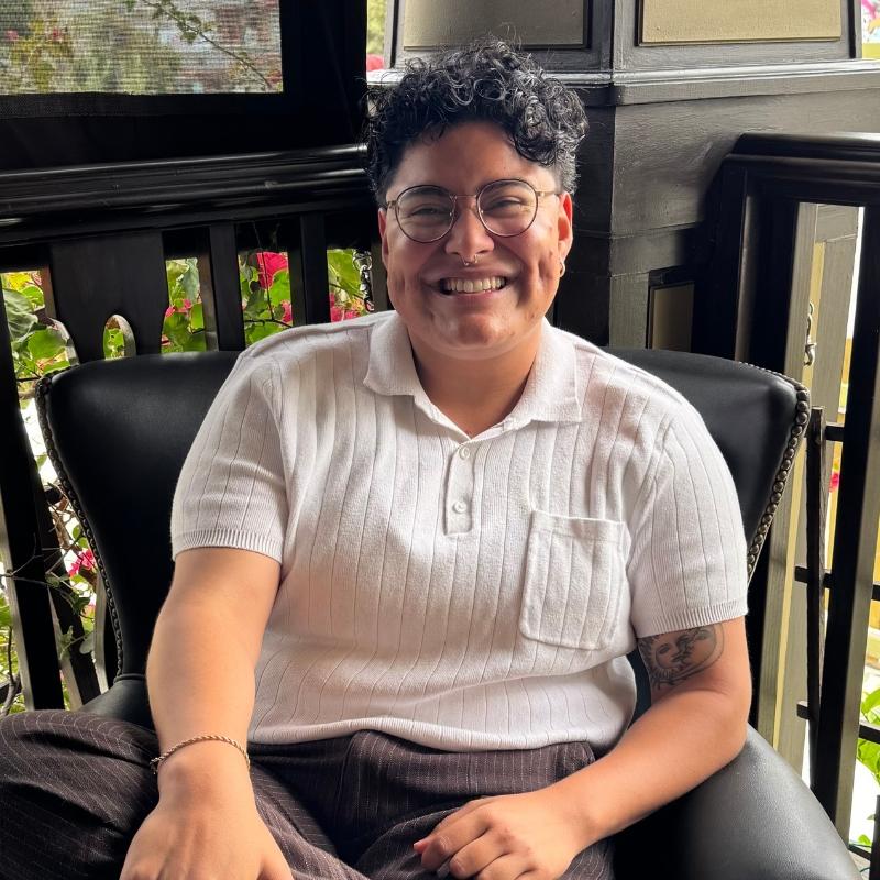 A photo of Gael Jerez-Urquia, LGBTQ CRC Program Coordinator. He is sitting on a chair in a corner in a white shirt. He has short black hair and glasses.